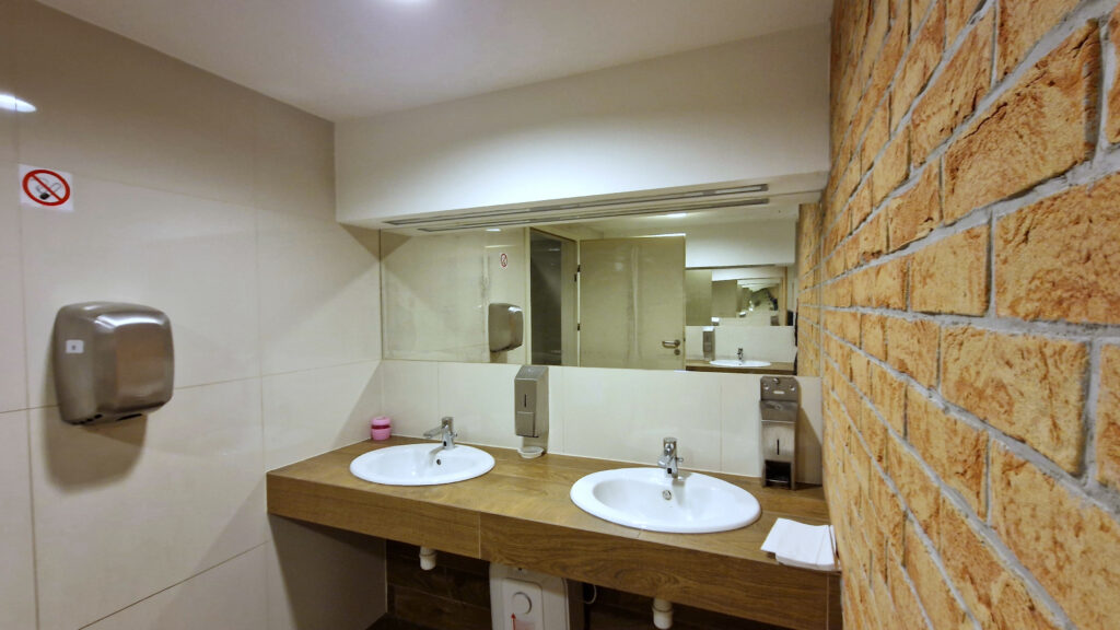 Strefa umywalek w miejskiej toalecie, wyposażona w dwa stanowiska z automatycznymi dozownikami mydła i suszarką do rąk. Płaski blat umożliwia łatwiejszy dostęp osobom z ograniczoną mobilnością. Jasne oświetlenie i lustro na całej szerokości zapewniają komfort użytkowania.