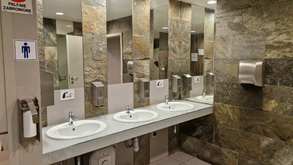Strefa umywalek w toalecie miejskiej utrzymana w wysokim standardzie czystości i estetyki. Trzy stanowiska z dozownikami mydła oraz suszarkami do rąk wkomponowane są w nowoczesne wnętrze, wykończone płytkami imitującymi kamień. Duże lustra, jasne oświetlenie oraz przejrzysta organizacja przestrzeni sprzyjają komfortowi użytkowników.