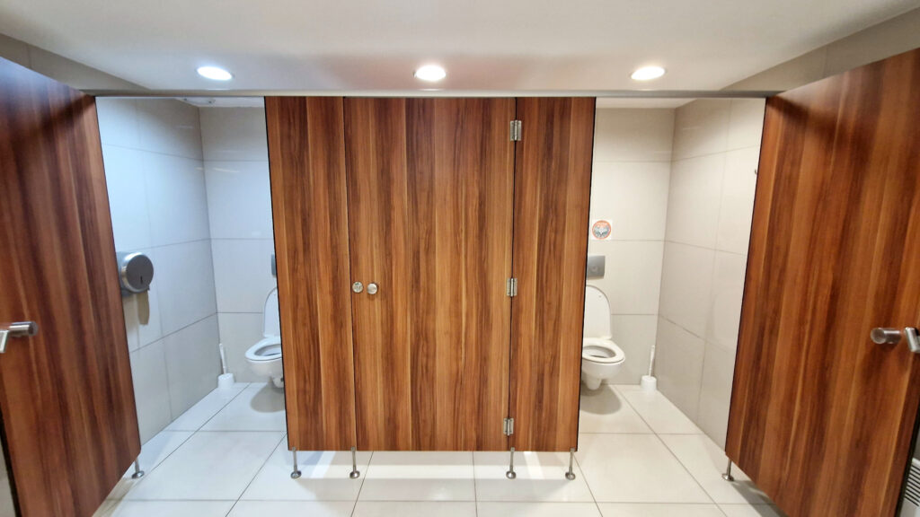 Strefa toalet zamykanych w miejskiej toalecie. Każda kabina zapewnia prywatność i wygodę, a stylizowane na drewno drzwi w połączeniu z jasnymi flizami i podłogą podkreślają estetykę oraz łatwość utrzymania czystości. Przestrzeń jest dobrze oświetlona i uporządkowana, co zwiększa komfort użytkowania.