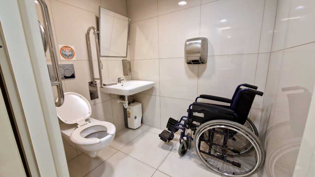 Toaleta miejska dostosowana do potrzeb osób z niepełnosprawnościami. Pomieszczenie wyposażone jest w poręcze ułatwiające korzystanie z toalety, przestrzeń do manewrowania wózkiem oraz umywalkę zamontowaną na odpowiedniej wysokości. Obok umywalki znajduje się suszarka do rąk, dozownik mydła i lustro ustawione pod kątem. W pomieszczeniu umieszczony jest wózek inwalidzki, co dodatkowo podkreśla jego dostępność dla osób o ograniczonej mobilności.