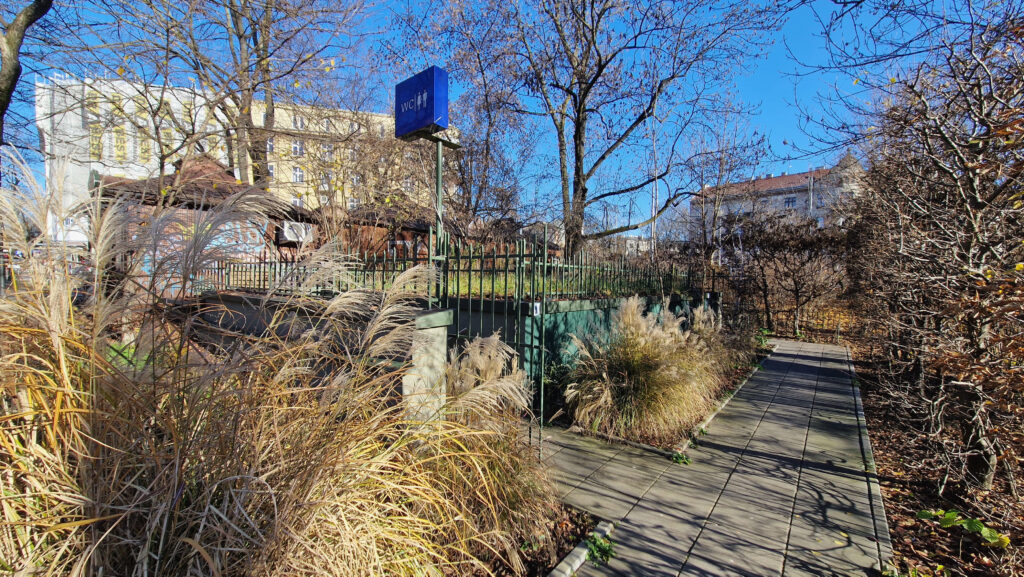 Wejście do miejskiej toalety umiejscowione wśród roślinności w Parku Krakowskim. Do obiektu prowadzi utwardzona, równa ścieżka zapewniająca wygodny dostęp osobom z ograniczoną mobilnością. Otoczenie jest zadbane, a wyraźne oznakowanie ułatwia znalezienie toalety.