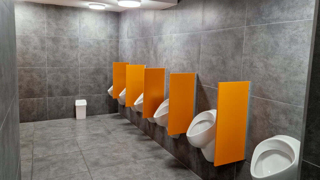 Strefa pisuarów w toalecie miejskiej, utrzymana w bardzo wysokim standardzie czystości. Nowoczesne wykończenie ścian grafitowymi płytkami podkreśla estetykę i funkcjonalność przestrzeni. Każdy pisuar oddzielony jest wygodną przegrodą zapewniającą prywatność użytkownikom, a oświetlenie LED dopełnia efekt schludności i porządku.