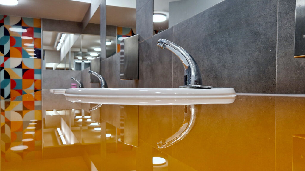 Umywalka w miejskiej toalecie, na tle nowoczesnej, kolorowej ściany oraz eleganckich, grafitowych płytek. Estetyczny blat o wysokim połysku podkreśla dbałość o czystość pomieszczenia. Nowoczesne baterie i dozowniki mydła wyposażone w czujniki zbliżeniowe są łatwo dostępne dla wszystkich użytkowników