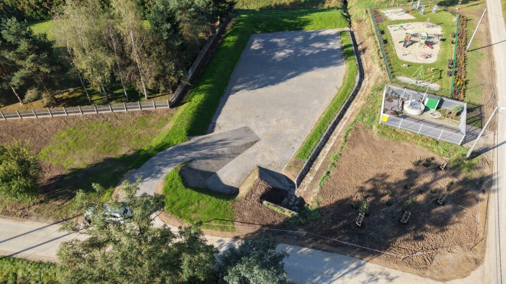Widok z lotu ptaka na nowo wybudowaną infrastrukturę hydrotechniczną w sąsiedztwie terenów rekreacyjnych. W centralnej części znajduje się duży zbiornik retencyjny o dnie wyłożonym szarymi płytami ażurowymi, otoczony trawiastymi skarpami. Obok zbiornika widoczny jest ogrodzony plac zabaw z kolorowymi urządzeniami oraz ogrodzona stacja techniczna z elementami aparatury pomiarowej. Na pierwszym planie widać obszar z nowymi nasadzeniami młodych drzew zabezpieczonych palikami oraz drogę dojazdową. Całość otoczona jest zielenią i drzewami liściastymi.