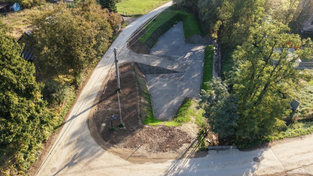 Widok z lotu ptaka na nowo wybudowany zbiornik retencyjny zlokalizowany na zakręcie drogi gruntowej w terenie podmiejskim. Zbiornik ma nieregularny, geometryczny kształt, a jego dno oraz nachylone skarpy wyłożone są szarymi ażurowymi płytami betonowymi, które umożliwiają infiltrację wód opadowych. Całość otoczona jest pasem zieleni i wysokimi, gęstymi drzewami. W centralnej części widoczna jest skarpa wjazdowa, umożliwiająca dostęp do wnętrza zbiornika.