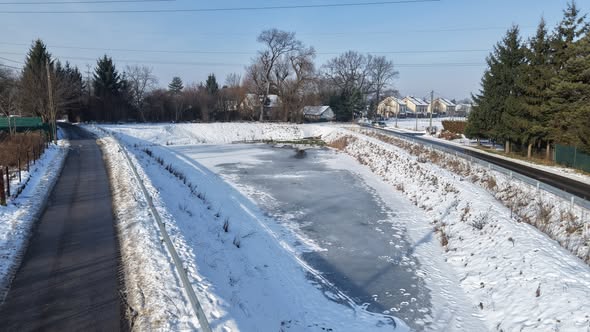 Zdjęcie przedstawia otwarty zbiornik retencyjny w warunkach zimowych, z lustrem wody pokrytym warstwą lodu i śniegu. Zbiornik znajduje się w obniżeniu terenu, a jego strome skarpy są całkowicie przykryte śniegiem. Po lewej stronie, wzdłuż krawędzi zbiornika, biegnie asfaltowa droga lub ścieżka oddzielona barierką energochłonną. W tle widoczna jest niska zabudowa mieszkaniowa, drzewa iglaste oraz linia horyzontu pod czystym niebem. Całość dokumentuje funkcjonowanie infrastruktury hydrotechnicznej ZIW w okresie zimowym.